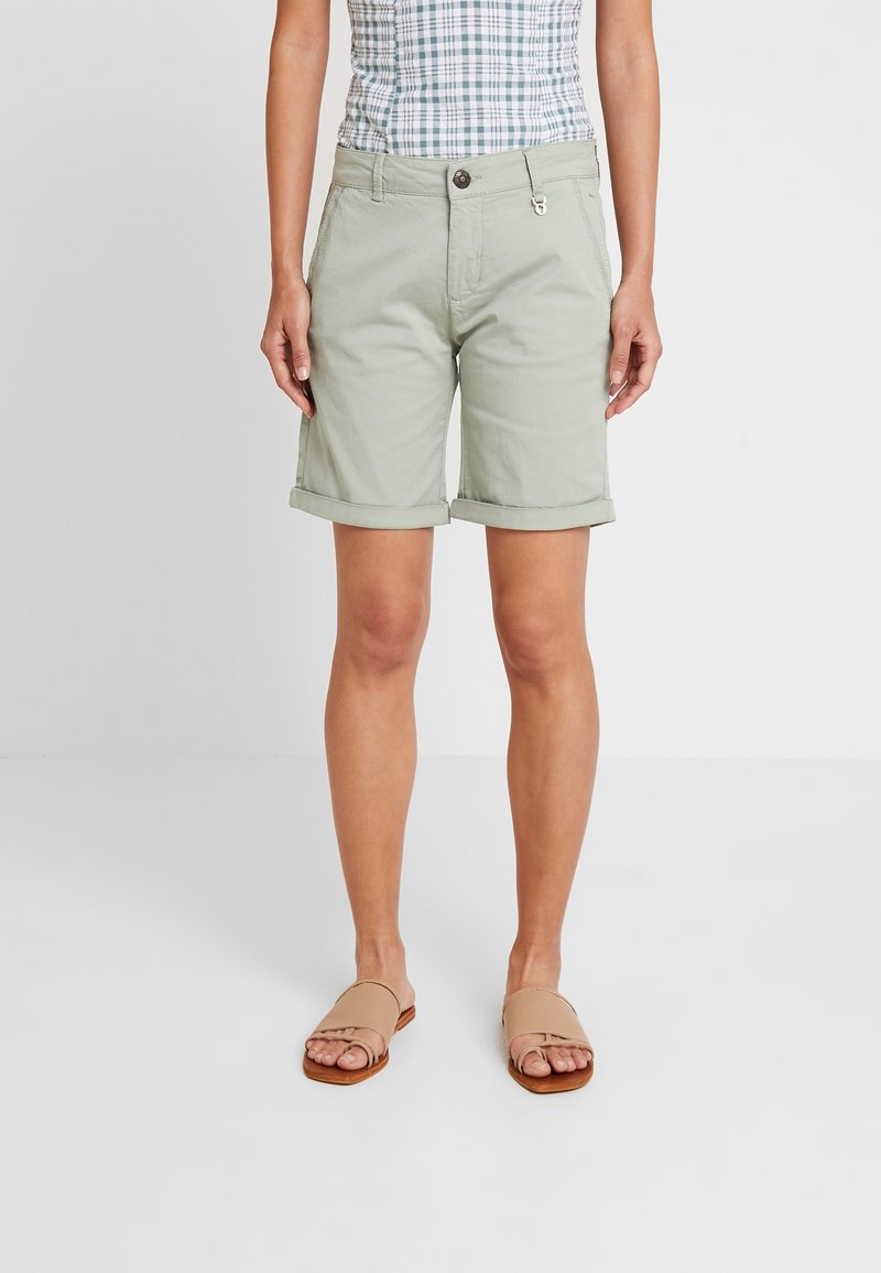 Světle zelené bavlněné šortky s ohrnutými lemy, vybavené zapínáním na knoflíky a kapsami, v kombinaci s béžovými nazouváky.