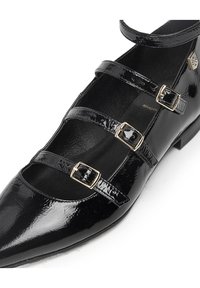 Zapato plano de charol negro con tres correas ajustables, puntera afilada y hebillas en tono dorado. Textura suave y tacón bajo.