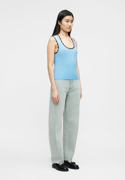 Γυναίκα που φοράει ανοιχτό γαλάζιο αμάνικο τοπ με μαύρη επένδυση, φαρδιά τζιν με ανοιχτό πλύσιμο και μαύρες μοκασίνες, στέκεται μπροστά σε απλό φόντο.