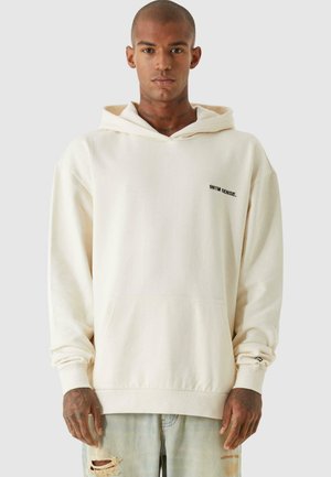 Cremiger Hoodies aus weichem Stoff, mit einer Kängurutasche, gerippten Bündchen und einem kleinen schwarzen Logo auf der linken Brust.