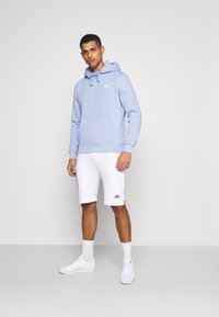 Felpa in cotone azzurro chiaro con tasca frontale e coulisse, abbinata a pantaloni corti in cotone bianchi con stampa del logo laterale e sneakers bianche.