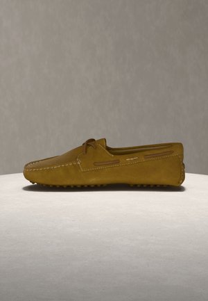 Bruine suède leren rijloafer met moc-toenaad, veterdetail en rubberen nopzool op een lichtgrijze ondergrond.