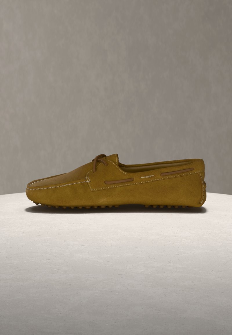 Καφέ μοκασίνι οδήγησης από καστόρι με ραφές moc toe, λεπτομέρεια με κορδόνι και σόλα από καουτσούκ με εξογκώματα σε ανοιχτό γκρι επιφάνεια.