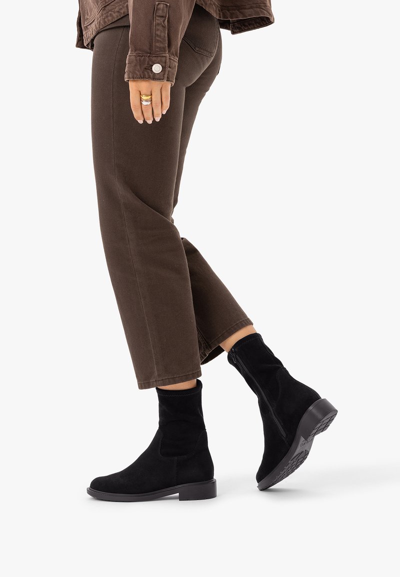 Personne portant un pantalon marron et des bottines noires, marchant avec un pied légèrement levé, visible de mi-cuisse jusqu'en bas sur fond blanc.