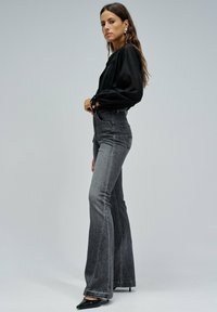 Kobieta stojąca bokiem, ubrana w czarną bluzkę z długimi rękawami, szare dżinsy w stylu dzwonów z wysokim stanem, czarne szpilki i srebrne kolczyki.