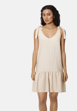 Mujer con cabello rizado que lleva un vestido beige sin mangas con tirantes de lazo y un dobladillo con volantes, de pie frente a un fondo liso.