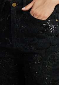 Pantaloni in denim nero con ricami in paillettes floreali, dalla forma aderente e una tasca frontale. La texture è leggermente lucida con dettagli intricati.