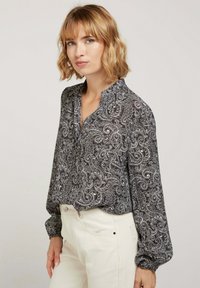 Blouse à motif paisley en noir et blanc avec un col popover et de longues manches froncées, associée à un pantalon de couleur crème.