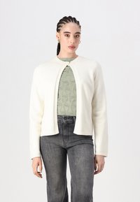 Cardigã branco justo com decote redondo, fecho de botão e bordas caneladas, usado sobre uma blusa verde texturizada e jeans escuros.