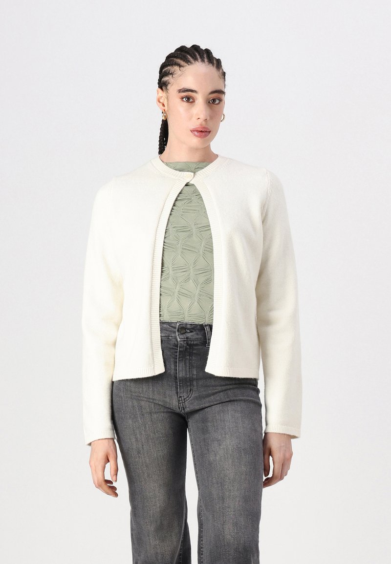 Cardigã branco justo com decote redondo, fecho de botão e bordas caneladas, usado sobre uma blusa verde texturizada e jeans escuros.