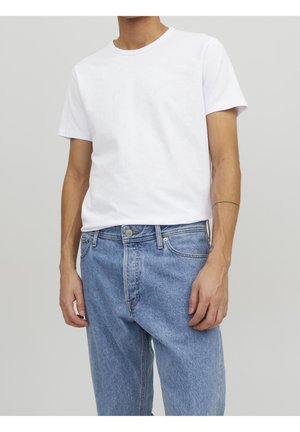 Homme portant un T-shirt blanc uni à manches courtes, rentré dans un jean en denim bleu clair taille haute, se tenant devant un fond neutre.