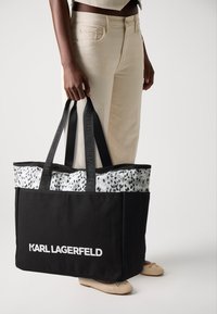 Svart tote-veske med grå og hvit dyremønsterkant, utstyrt med to svarte håndtak med preget "KARL LAGERFELD" merke.