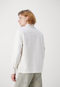 Chaqueta de mezclilla blanca con un corte relajado, puños abotonados y detalles de costura sutiles. Combinada con pantalones de un color claro.