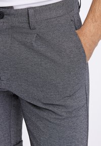 Grau strukturierte Hose mit dezentem Muster, ausgestattet mit Knopfverschluss und Seitentaschen. Das Material wirkt weich und die Passform ist maßgeschneidert.