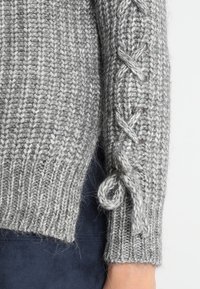 Grauer Strickpullover mit strukturiertem Muster, dekorativen geflochtenen Akzenten an den Ärmeln und gebundenen Details an den Bündchen.