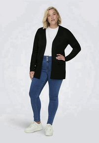 Cardigan longo preto com abertura frontal, usado sobre uma blusa branca. Combinado com jeans skinny azuis e ténis brancos. Design minimalista e corte casual.