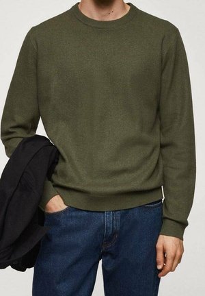 Mand iført olivengrøn striktrøje og blå jeans, der holder en sort jakke i den ene hånd.
