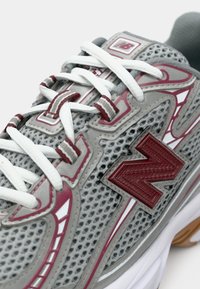 Chaussure de sport grise avec tige en mesh, accents rouges et blancs, dotée d'un logo rouge, de lacets blancs et d'une semelle en caoutchouc texturée.