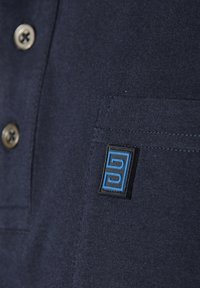 Navy polo shirt with a smooth texture, featuring round gold buttons and a square blue logo patch on the left side.