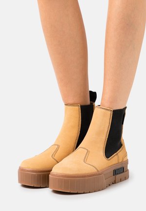 Bottines en suède beige avec des panneaux latéraux élastiques noirs, une épaisse semelle en caoutchouc marron et des détails de couture prononcés. Tirant noir à l'arrière.
