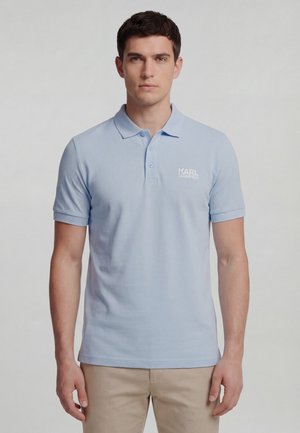 Młody mężczyzna ubrany w jasnoniebieską koszulkę polo Karla Lagerfelda i beżowe spodnie, stojący na jednolitym szarym tle.