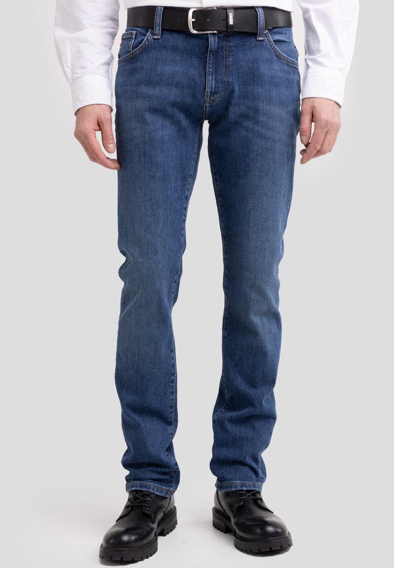 Férfi kék slim-fit farmert, fekete övet, fehér inget és fekete bőr csizmát viselve, egyszínű háttér előtt áll.