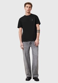 T-shirt di cotone nera con colletto rotondo e piccolo logo, abbinata a jeans grigi chiari a gamba larga. Indossata con scarpe nere. Sfondo neutro.