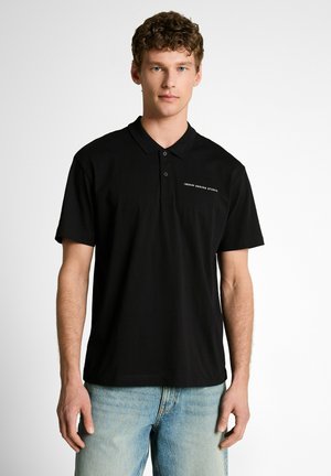 Young man with curly hair wearing a black polo shirt with "Denim Design Studio" text, paired with light blue jeans, standing against white background.