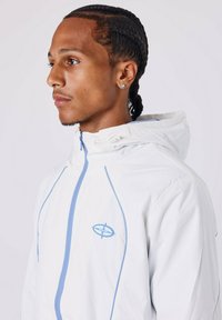 Jonge man met gevlochten haar die een witte hoodie met lichtblauwe rits en logo draagt, kijkt naar links tegen een eenvoudige achtergrond.