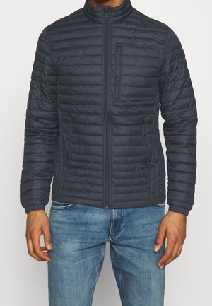 Mężczyzna ubrany w granatową pikowaną kurtkę na suwak z dwoma bocznymi kieszeniami i jedną zapinaną na suwak kieszenią na piersi, w połączeniu z jasnoniebieskimi dżinsami.