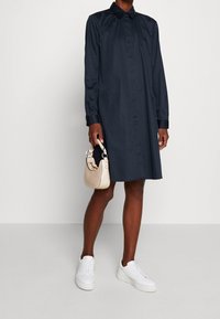 Tmavě modré šaty ve stylu košile z hladkého materiálu, s knoflíkovým zapínáním vpředu, límečkem, dlouhými rukávy a uvolněným střihem. V kombinaci s bílými teniskami a malou světlou kabelkou.