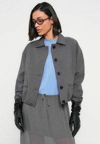 Γυναίκα με ίσια μαύρα μαλλιά και γυαλιά, φορά γκρι σακάκι με κουμπιά, ανοιχτό μπλε πλισέ τοπ, γκρι φούστα και μαύρα δερμάτινα γάντια.