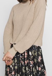 Beige gestrickter Pullover mit hohem Kragen und gerippter Textur, kombiniert mit einem schwarzen, blumigen Plisseerock mit bunten botanischen Mustern.