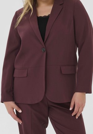 Kvinde iført en mørk burgunder blazer med en knap og matchende bukser, sort top under, stående med hænderne afslappet ned langs siderne.