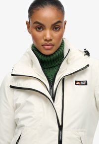 Biała wodoodporna kurtka z czarnymi zamkami błyskawicznymi, wyposażona w wysoki kołnierz i naszywkę z logo. Nosi się ją na ciemnozielony dziany golf.