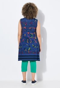 Sukienka bez rękawów w kolorze niebieskim z kolorowymi geometrycznymi wzorami, zestawiona z zielonymi legginsami capri. Modelka nosi granatowe buty wsuwane.