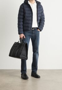 Navy puffer jacket, white polo shirt, dark jeans, black leather shoes. Holding a large black tote bag with a textured surface and dual handles.