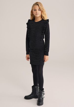 Schwarzes Langarmkleid mit Rüschendetails, glitzernder Textur und figurbetonter Silhouette. Kombiniert mit schwarzen Knöchelstiefeln und Leggings.