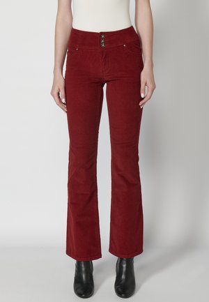 Femme portant un pantalon flare en velours côtelé rouge taille haute avec trois boutons et des bottines noires, debout sur un fond uni.