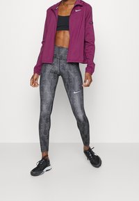 Coupe-vent Nike bordeaux, associé à un legging noir avec un logo et un texte imprimés en blanc. Chaussures de sport noires avec des accents gris.