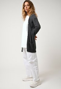 Kolvgrå cardigan med långa ärmar, framknappar och fickor, bärs över en vit skjorta och tillsammans med lösa vita byxor och beiga sneakers.