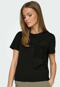 Svart bomullströja med rund halsringning, korta ärmar och en texturerad bröstficka med ett cirkulärt mönster på vänster bröst.