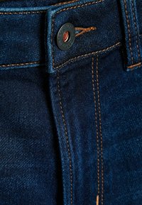 Donkerblauwe denimstof met contrasterende oranje stiksels, een metalen knoopsluiting en een subtiele textuur zonder zichtbare patronen.