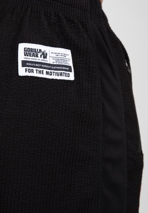 Zwarte trainingsshorts van een gestructureerde stof, met een opvallend wit label met zwarte tekst en logo, dat de materiaalsamenstelling en branding toont.