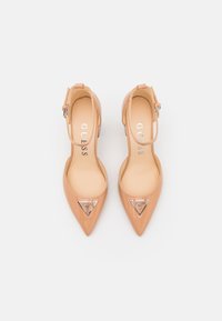 Scarpe con tacco alto in vernice color carne, con punta affusolata, cinturini alla caviglia e un logo triangolare argento sul davanti. Texture liscia.
