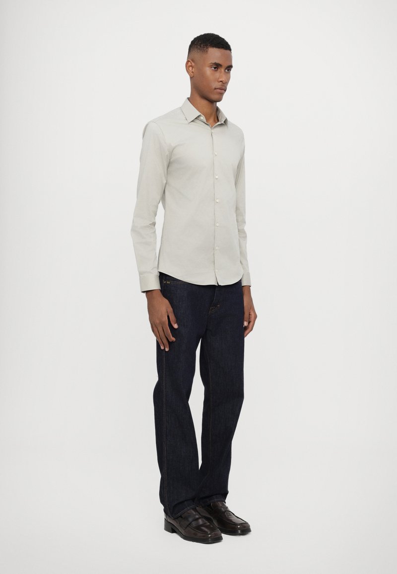 Chemise button-up gris clair à manches longues, associée à un jean droit bleu foncé et des chaussures noires. Tissu lisse, design simple.