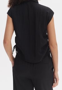 Černý top bez rukávů s límečkem, texturovaným materiálem a nastavitelnými šňůrkami na bocích, v kombinaci s černými kalhotami s detailem zadní kapsy.