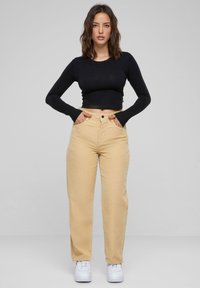 Top corto nero a maniche lunghe abbinato a pantaloni in corda beige a vita alta. Sneakers bianche completano l'outfit. Sfondo neutro.