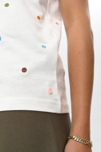 Nahaufnahme einer Person, die ein weißes Hemd mit bunten gestickten Punkten und olivgrüne gerippte Hosen trägt, und ein goldenes Armband am Handgelenk zeigt.