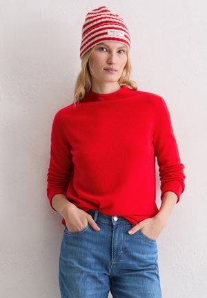 Un pull en laine rouge avec un col rond et des manches longues, associé à un jean en denim bleu. Un bonnet tricoté rouge et blanc rayé avec une étiquette.